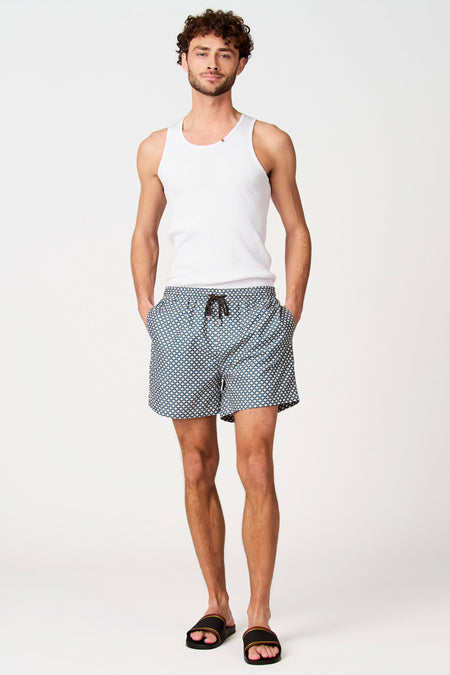Short de bain bleu-vert à motif géométrique blanc de Bruce & Butler, porté par un homme avec une chemise blanche et des sandales rayées.