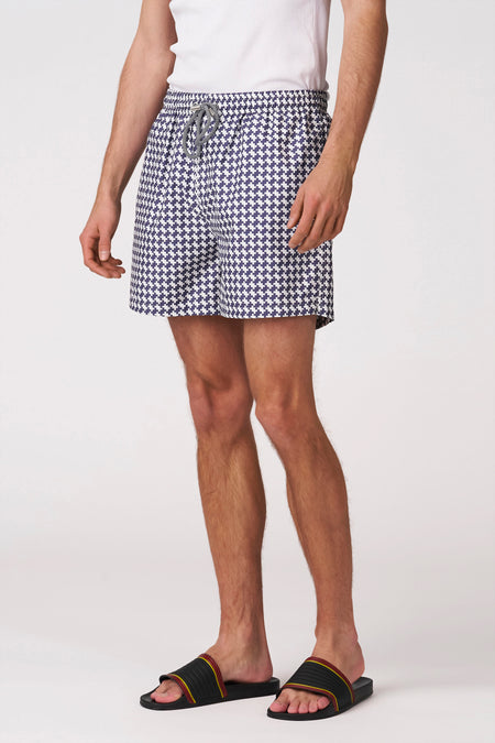 Short de bain pied-de-poule bleu et blanc de Bruce & Butler, porté par un homme de dos avec un débardeur blanc et des sandales noires.