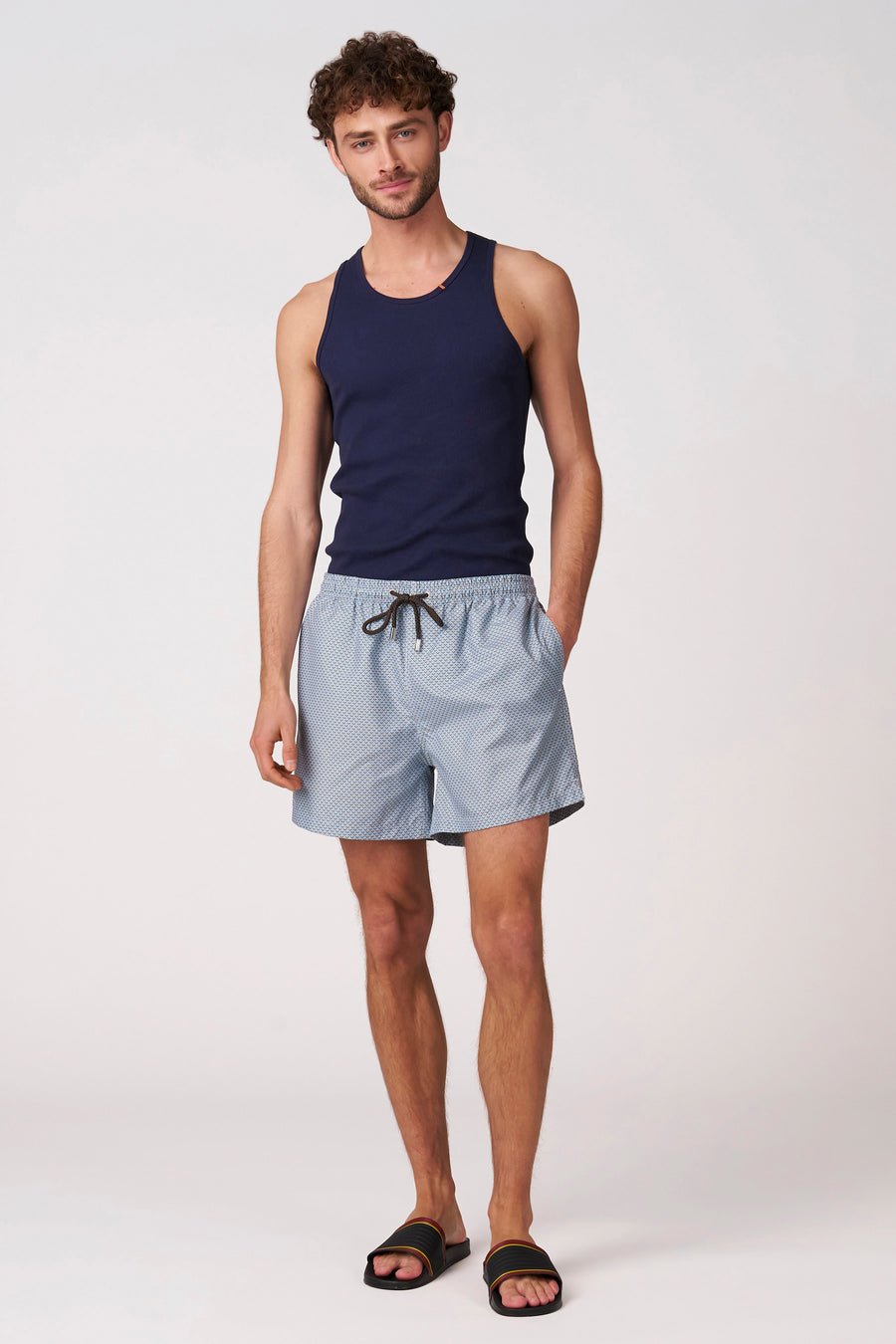 Short de bain bleu de Bruce & Butler avec motif géométrique bleu clair, porté par un homme en débardeur bleu foncé et tongs noires.