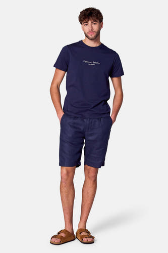 Blauw T-shirt met korte mouwen van Cyclo Club Marcel, met witte tekstprint, gecombineerd met blauwe linnen shorts en bruine sandalen.
