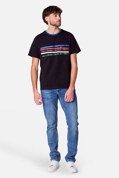 T-shirt noir à manches courtes de Cyclo Club Marcel, avec des rayures horizontales colorées et porté avec des jeans bleus et des baskets blanches.