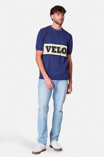 T-shirt bleu à manches courtes de Cyclo Club Marcel, avec le texte 'VELO' sur la poitrine, associé à des jeans et des baskets blanches.