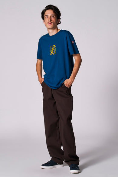 T-shirt bleu à manches courtes du Cyclo Club Marcel, avec une impression jaune et porté avec un pantalon marron et des baskets bleues.