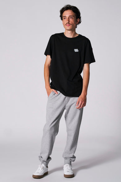T-shirt noir à manches courtes de Cyclo Club Marcel, avec petit logo, porté avec un short de jogging gris et des baskets blanches.