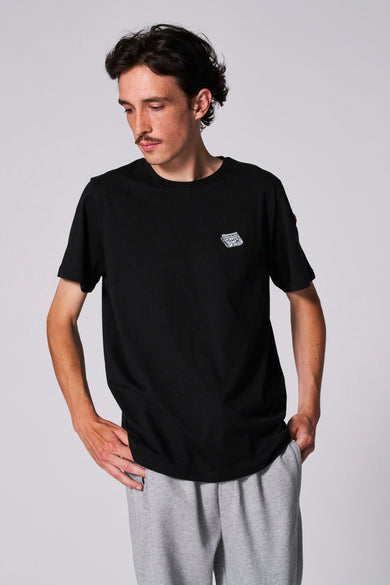 T-shirt noir à manches courtes de Cyclo Club Marcel, avec une petite impression d'un journal sur la poitrine, associé à un pantalon de jogging gris.