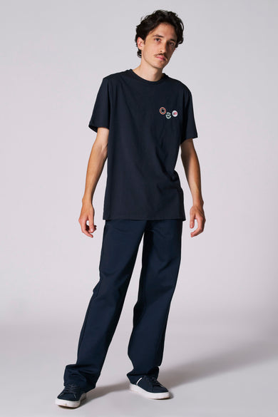 T-shirt bleu à manches courtes de Cyclo Club Marcel, avec des badges sur la poitrine, associé à un pantalon bleu marine et des baskets.