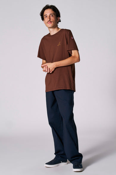 T-shirt brun à manches courtes du Cyclo Club Marcel, avec un petit logo et porté avec un pantalon bleu marine et des baskets.