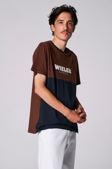 T-shirt bleu à manches courtes de Cyclo Club Marcel, avec un haut marron et des pantalons blancs.