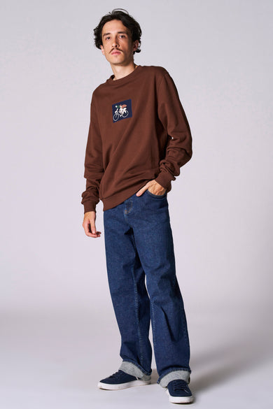 Pull-over marron du Cyclo Club Marcel, avec col rond et patch sur la poitrine, associé à des jeans et des baskets.