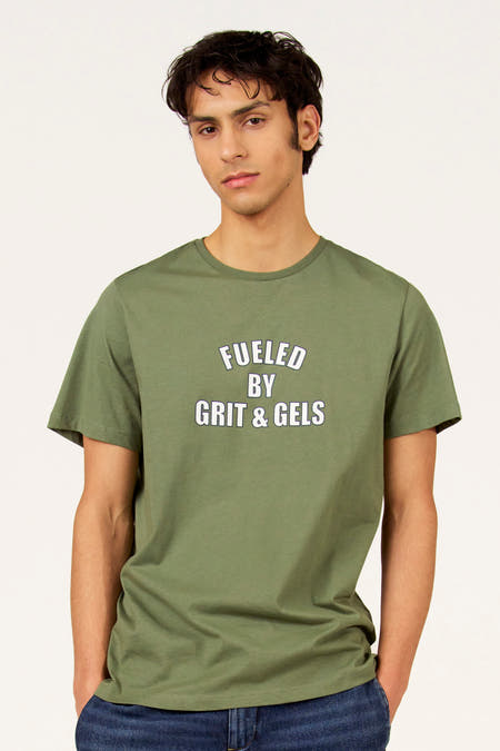 Groen T-shirt met korte mouwen van Cyclo Club Marcel, met de tekst "Fueled by Grit & Gels" en gedragen met een jeansbroek.
