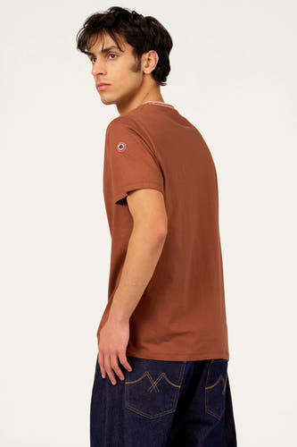 T-shirt marron à manches courtes de Cyclo Club Marcel, avec logo sur la manche et jean bleu foncé.