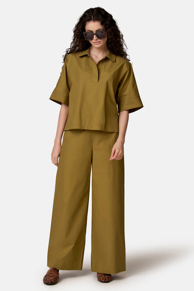 Pantalon vert de COPINES D'ANVERS à jambes larges, associé à un chemisier assorti, des lunettes de soleil et des mocassins à imprimé serpent.