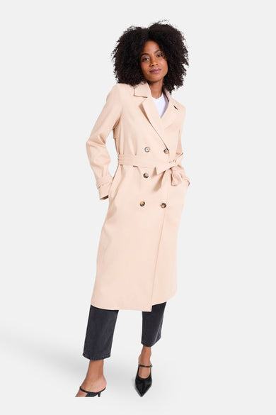 Manteau long beige de NAF NAF, avec une ceinture à nouer, associé à un pantalon noir et des bottes.