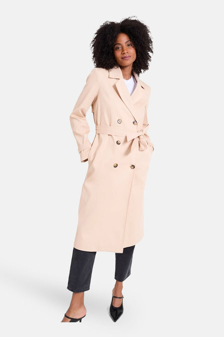 Manteau long beige de NAF NAF, avec une ceinture à nouer, associé à un pantalon noir et des bottes.