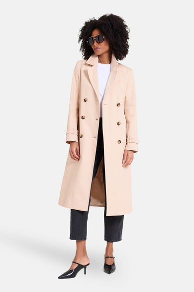 Manteau long beige de NAF NAF, avec une double rangée de boutons, porté avec un chemisier blanc et un jean noir.