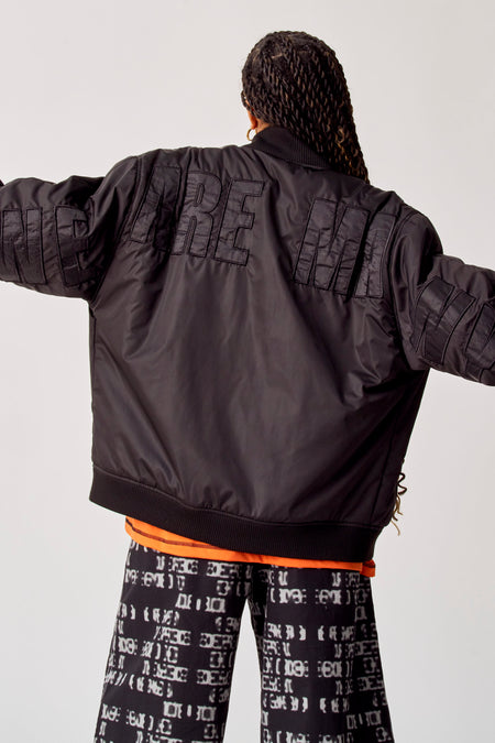 Manteau court noir de COELY avec du texte sur le dos, associé à une chemise orange et un pantalon imprimé.