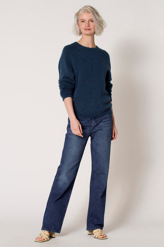 Vrouw met blauwe trui en jeans, beige sandalen, staand.
