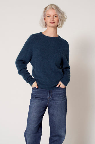 Vrouw met korte blonde haar in blauwe trui en jeans met handen in de zakken.