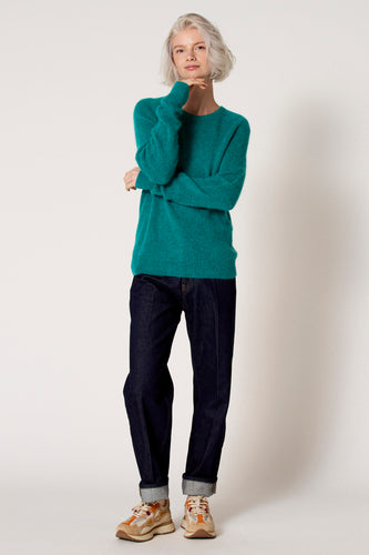 Vrouw met een turquoise trui, donkerblauwe jeans en kleurrijke sneakers, staand op een witte achtergrond.
