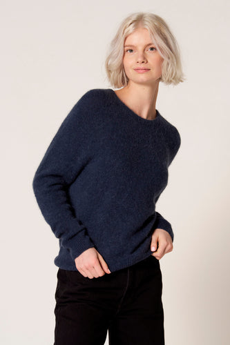 Vrouw in blauwe trui en zwarte broek voor een neutrale achtergrond.