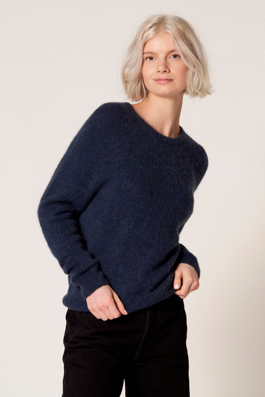 Vrouw in blauwe trui en zwarte broek voor een neutrale achtergrond.