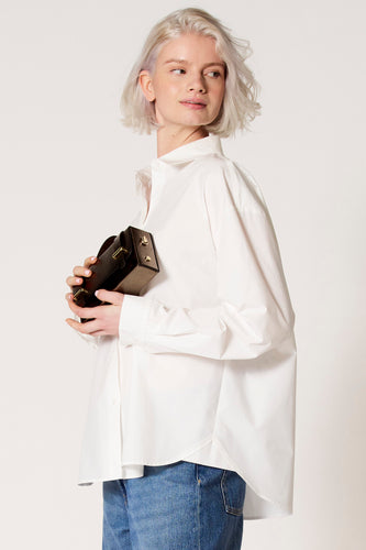 Vrouw met blond haar draagt witte blouse en blauwe jeans, houdt bruine tas vast, witte achtergrond.
