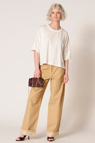 T-shirt écru à manches courtes de Clouds of Fashion, avec des manches amples et une subtile fente sur le côté, combiné avec un pantalon marron.