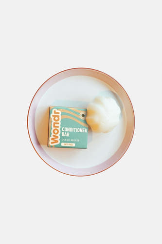 Barre de conditionneur blanche en forme de coquillage de WONDR CARE, à côté de l'emballage sarcelle avec des vagues orange, sur un plateau blanc avec un bord rose.