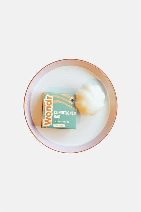 Barre de conditionneur blanche en forme de coquillage de WONDR CARE, à côté de l'emballage sarcelle avec des vagues orange, sur un plateau blanc avec un bord rose.