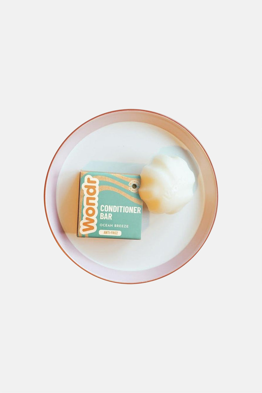 Barre de conditionneur blanche en forme de coquillage de WONDR CARE, à côté de l'emballage sarcelle avec des vagues orange, sur un plateau blanc avec un bord rose.