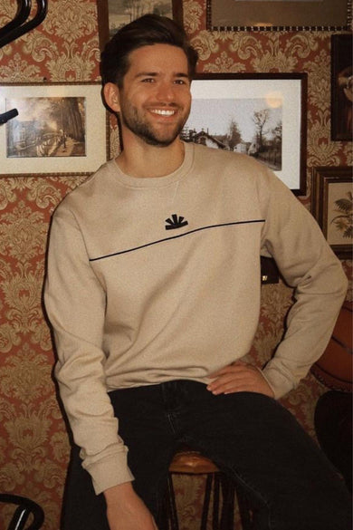 Beige sweater van BASELAB, met een zwarte streep en logo op de borst, gedragen met zwarte jeans.
