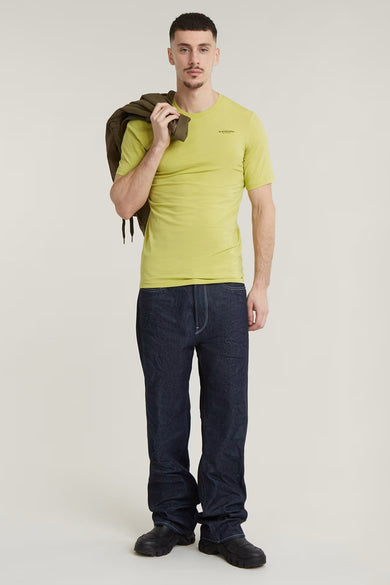 T-shirt jaune à manches courtes de G-Star RAW, porté avec un manteau sur l'épaule, associé à des jeans foncés et des chaussures noires.
