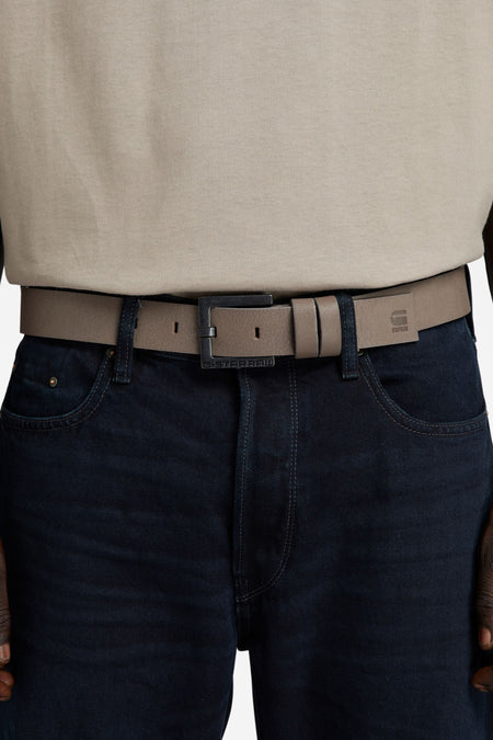Ceinture grise de G-Star RAW, portée avec un jean bleu foncé et une chemise beige.