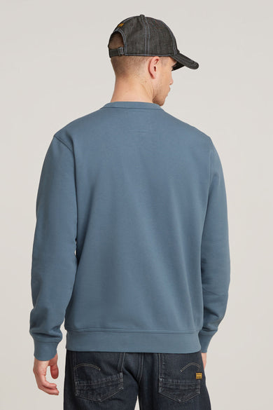 Sweater bleu à col rond de G‑Star RAW, vu de l’arrière, avec un jean sombre et une casquette en denim.