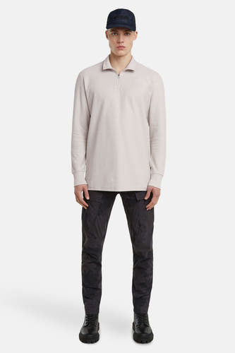 Gilet écru de G-Star RAW, avec des manches longues, porté avec un pantalon noir à imprimé camouflage et une casquette.