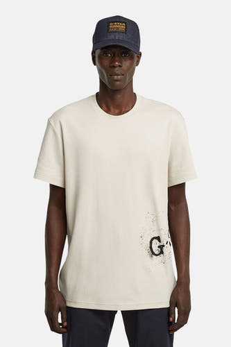 T-shirt écru à manches courtes de G-Star RAW, avec une impression sur l'arrière et porté avec un chapeau bleu marine.