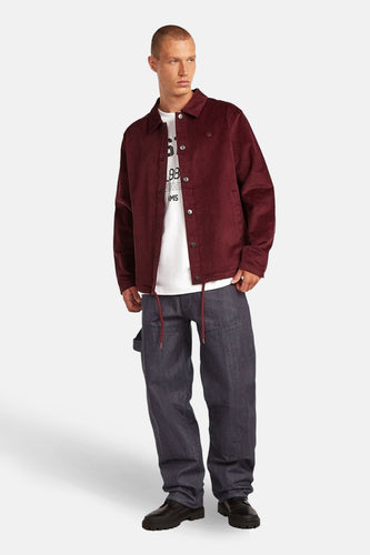 Rode jas van G-Star RAW, gedragen over een wit T-shirt en een losvallende donkerblauwe jeans met donkerbruine veters.