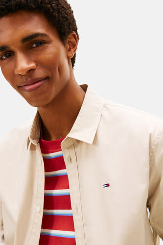 Chemise beige à longues manches de Tommy Jeans, avec un logo subtil et une chemise rouge rayée visible en dessous.