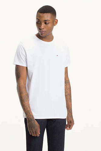 T-shirt blanc de TOMMY JEANS, à manches courtes, porté avec des jeans foncés et des tatouages sur les bras.