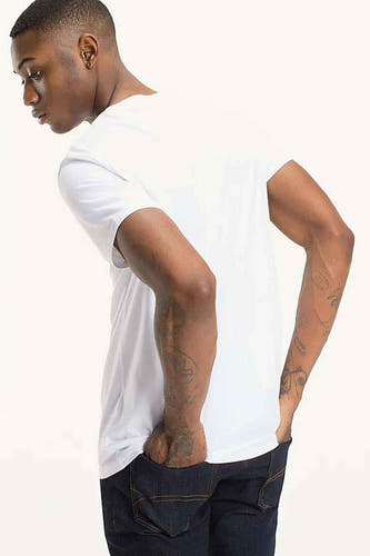 Wit T-shirt van TOMMY JEANS, met korte mouwen, gedragen met donkere jeans en tatoeages op de armen.