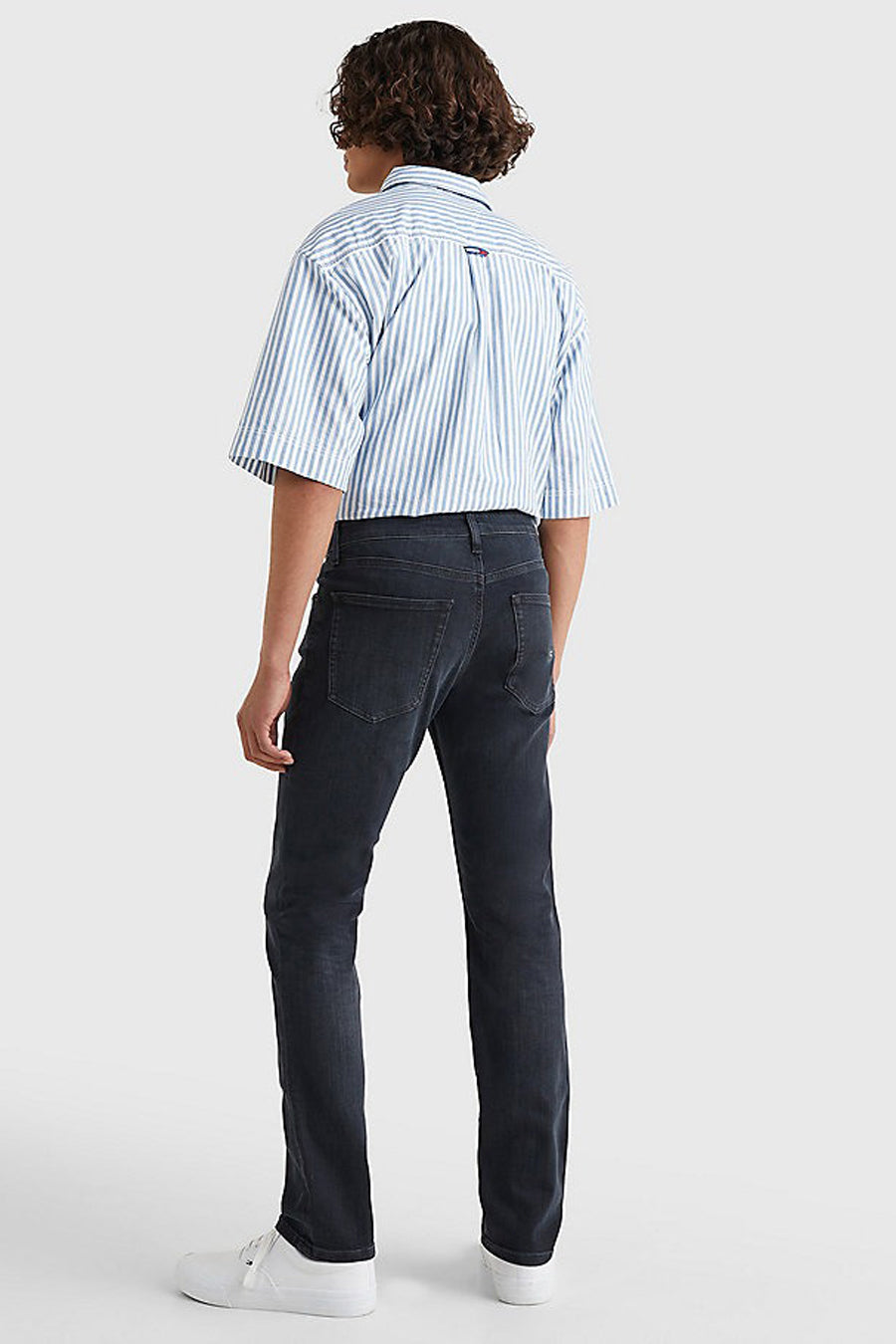 Donkergrijze slim fit jeans van TOMMY JEANS, gecombineerd met een blauw gestreept overhemd en witte sneakers, gezien van achteren.