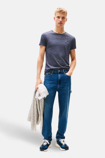 Man met blond haar in blauw T-shirt, jeans en blauwe sneakers, met een grijze trui in de hand, staand tegen een witte achtergrond.