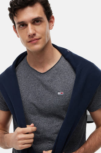 T-shirt gris chiné Tommy Jeans à col en V, petit logo sur la poitrine et un pull bleu marine sur les épaules.