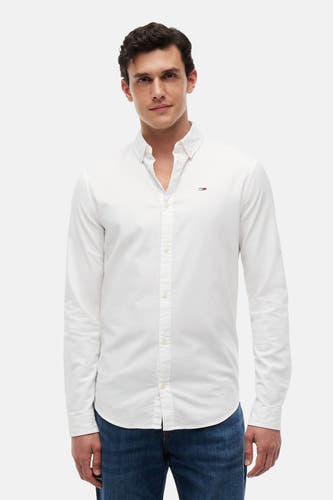 Chemise blanche à manches longues TOMMY JEANS, avec petit logo drapeau sur la poitrine, col boutonné, portée avec un jean bleu.