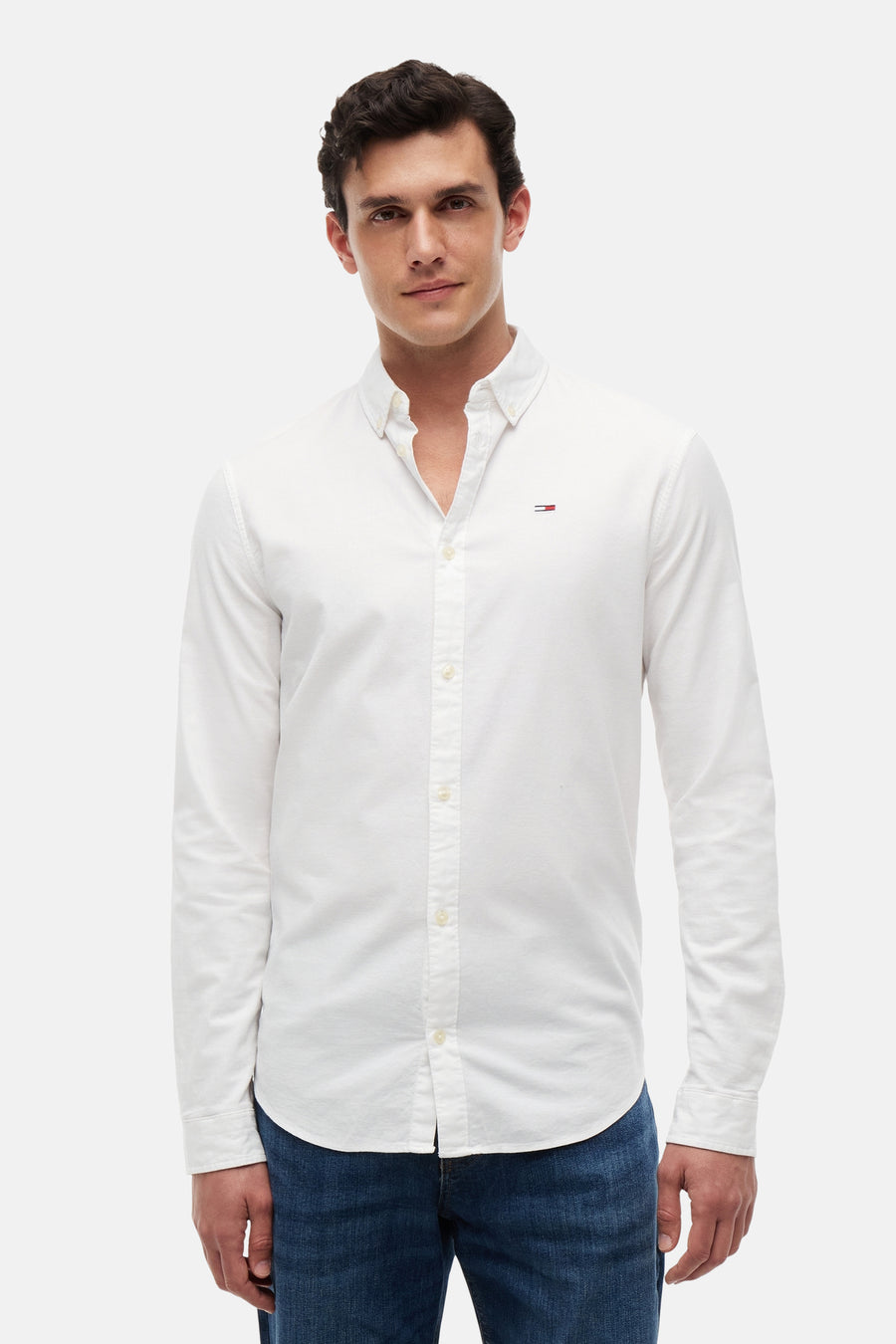 Chemise blanche à manches longues TOMMY JEANS, avec petit logo drapeau sur la poitrine, col boutonné, portée avec un jean bleu.