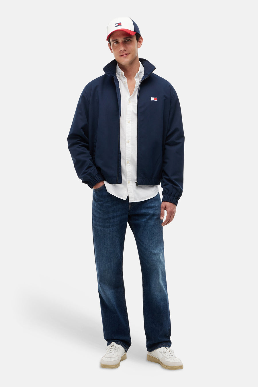 Chemise blanche à manches longues de TOMMY JEANS, portée sous une veste bleu marine, un jean et une casquette.