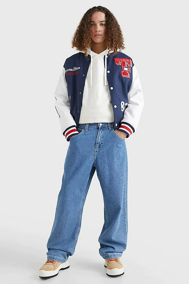Wijde jeans van midblauw denim van TOMMY JEANS, gedragen met een hoodie en een varsity jacket.