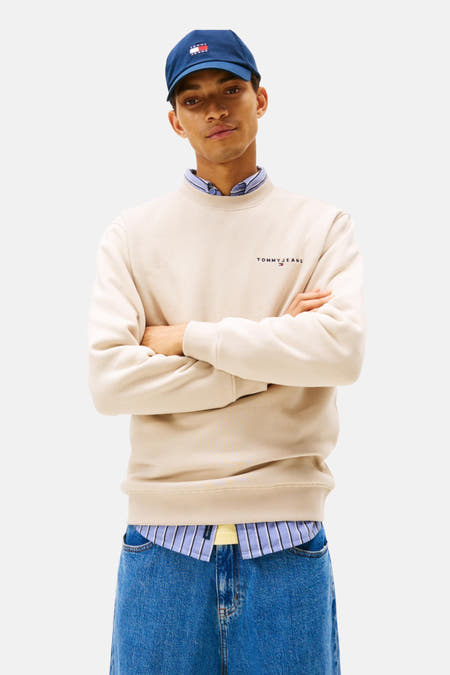 Gilet beige de TOMMY JEANS, avec des manches longues, porté avec des jeans bleus et un chapeau bleu marine.