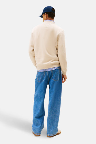 Man in beige trui, blauwe jeans en pet.