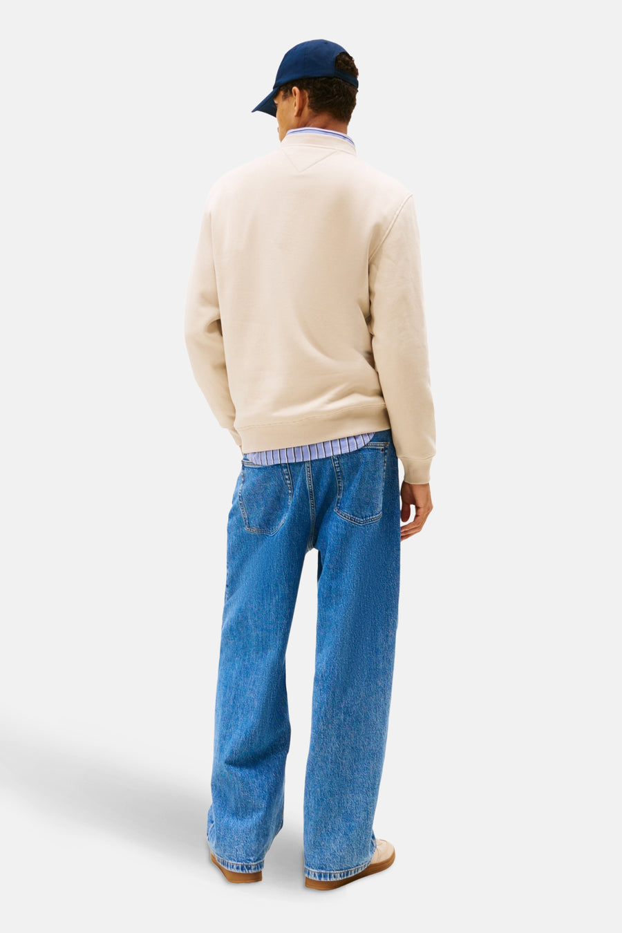 Man in beige trui, blauwe jeans en pet.